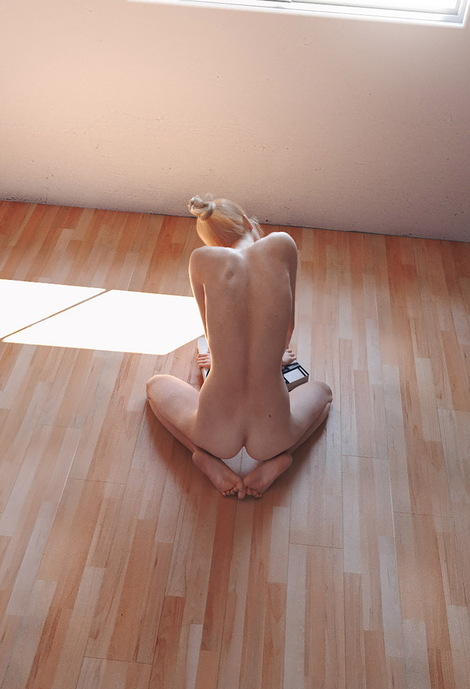 A woman with blonde hair tied in a bun sits nude on a wooden floor near a window with sunlight streaming in.